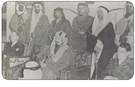 الملك عبد العزيزويظهر في الصورة من الخلف بعض ضباط الحرس الملكي وقوفاً من اليمين إلى اليسار العقيد / محمد عبد الله الذيب والفريق / سعيد جودت