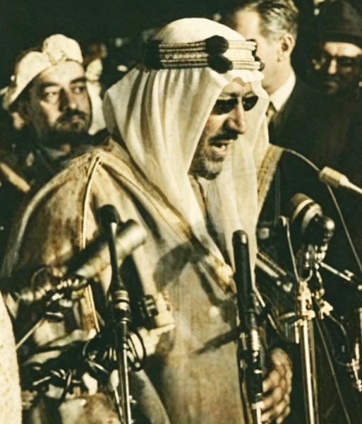 King Saud delivering a speech at a press conference in the United States, with Major General Abdulmunim Al‑Aqeel standing behind him, February 1957—may God have mercy on them.”