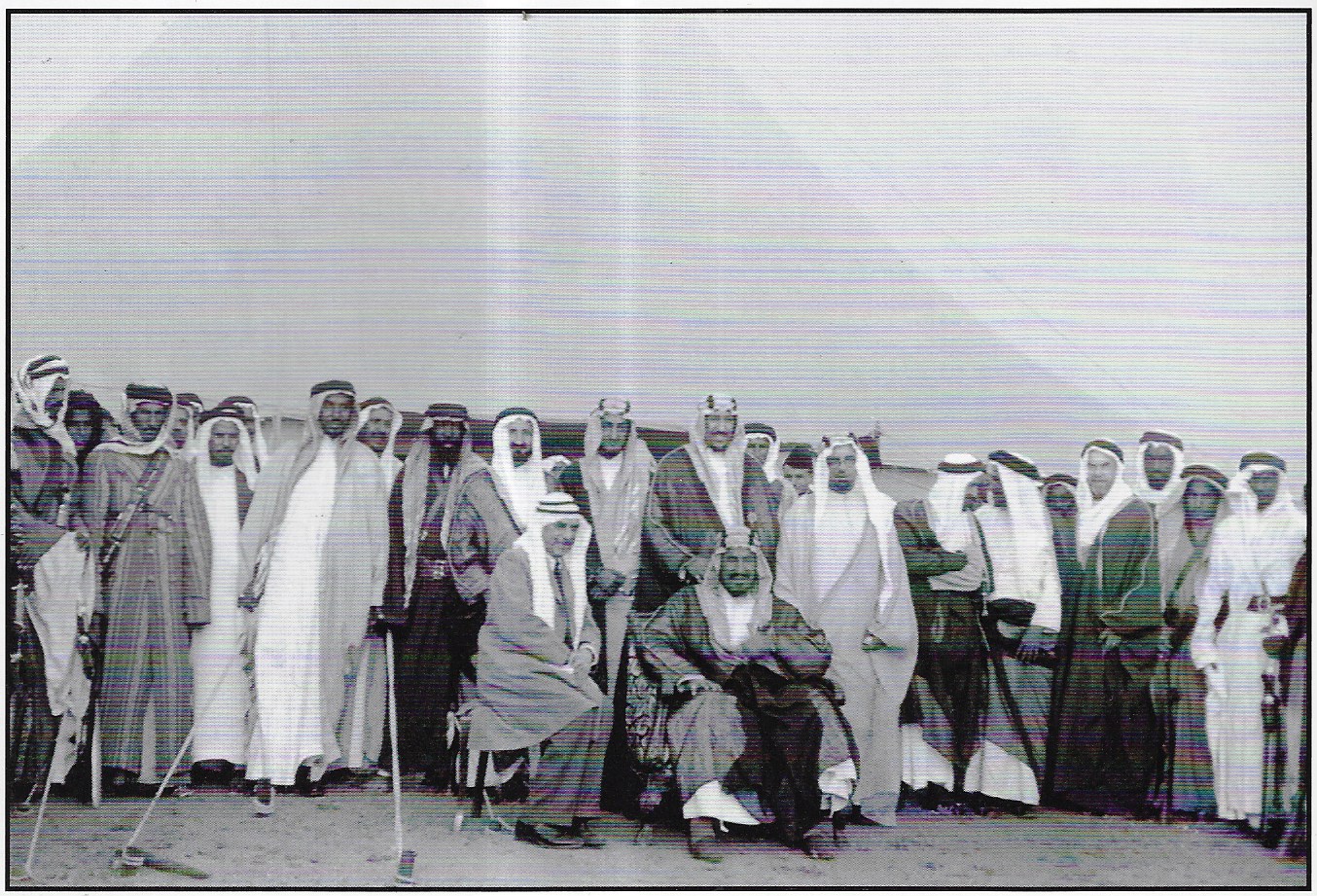 King Abdulaziz with a guest to his right, and behind them stand Princes Saud and Faisal (later kings), along with Yusuf Yassin, Abdulrahman Al‑Tubaishi, Hafiz Wahba, and Abdullah Philby
