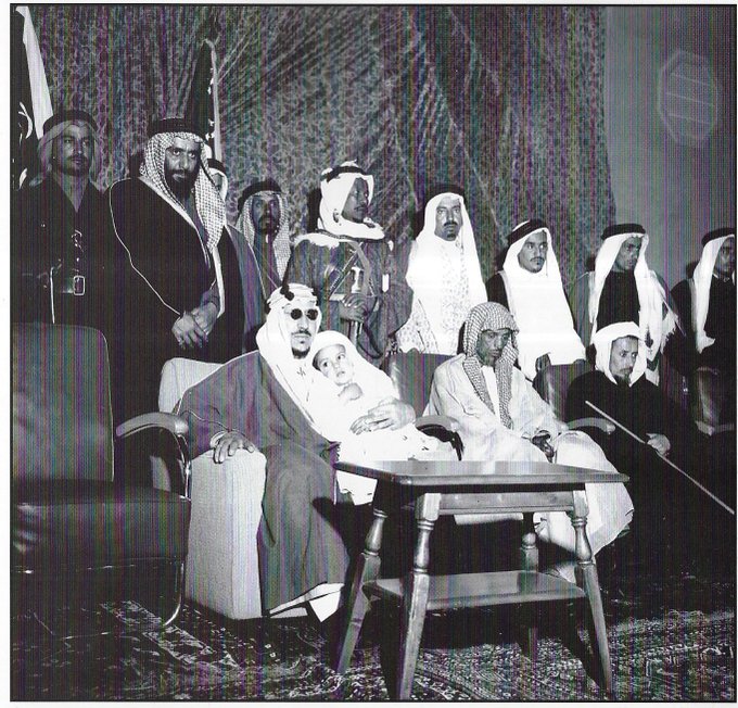 Crown Prince Saud (later King Saud) during a visit to the Eastern Province, holding his son Prince Mansour, with Prince Saud bin Jiluwi standing behind them.